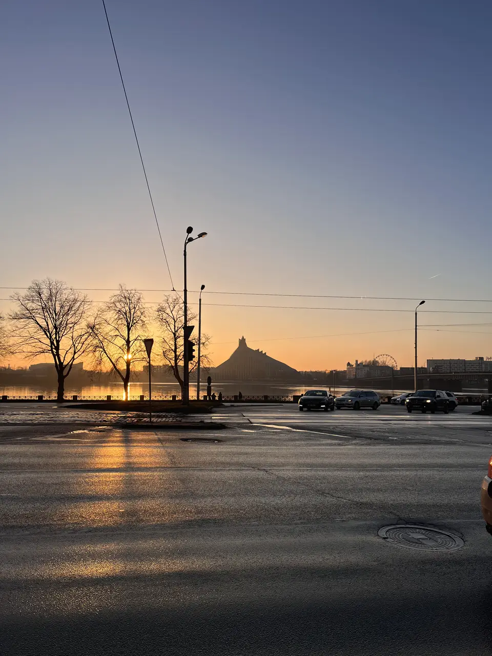 Low winter sun over a waterfront road, with bare trees, street lamps and wet tarmac reflecting the light