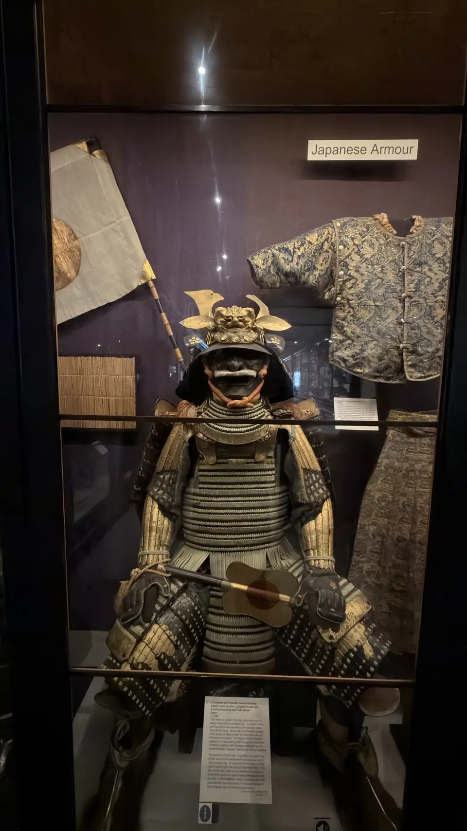 A suit of samurai armour displayed in a lit museum case beside a folding fan and embroidered garments